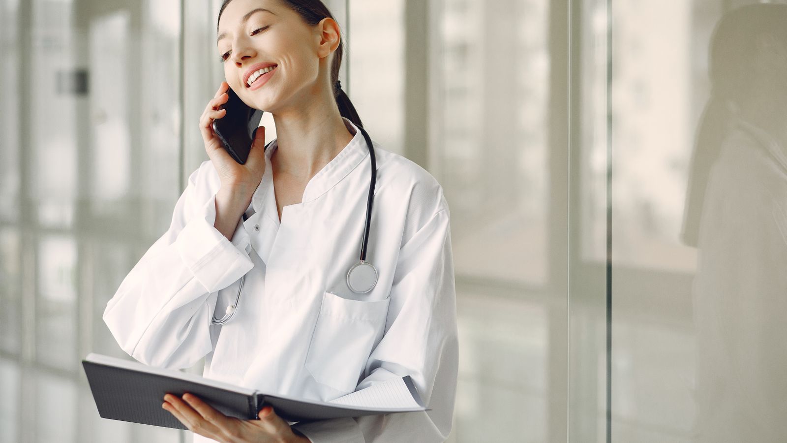 Smiling ethnic female doctor in medical uniform with stethoscope and notebook talking on smartphone while standing in glass hospital corridor