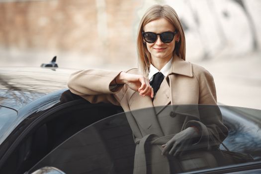 Smiling female driver in stylish coat and sunglasses leaning on automobile while standing on city street with paper bags