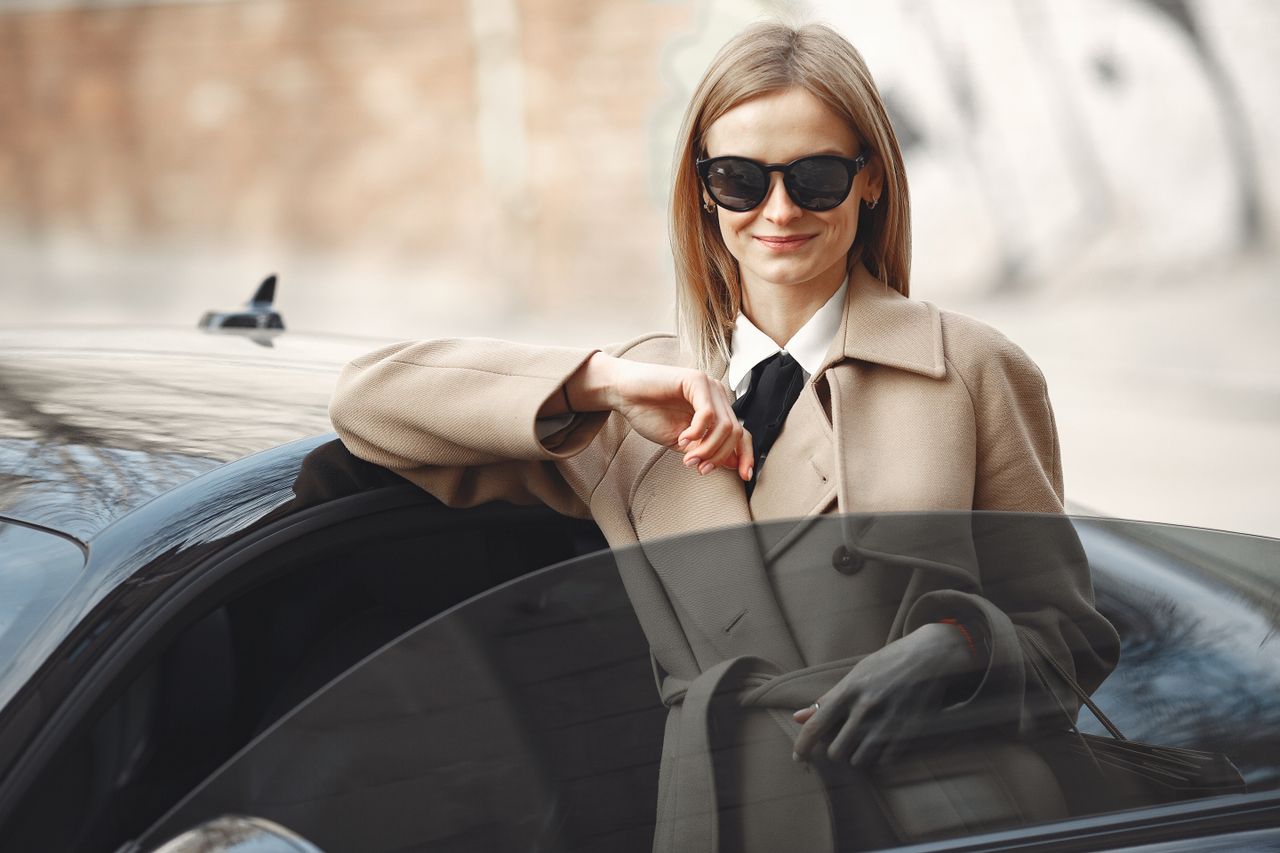 smiling female driver in stylish coat and sunglasses leaning on automo