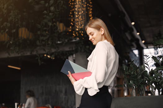 A stylish businesswoman stands in a contemporary cafe holding a colorful folder.