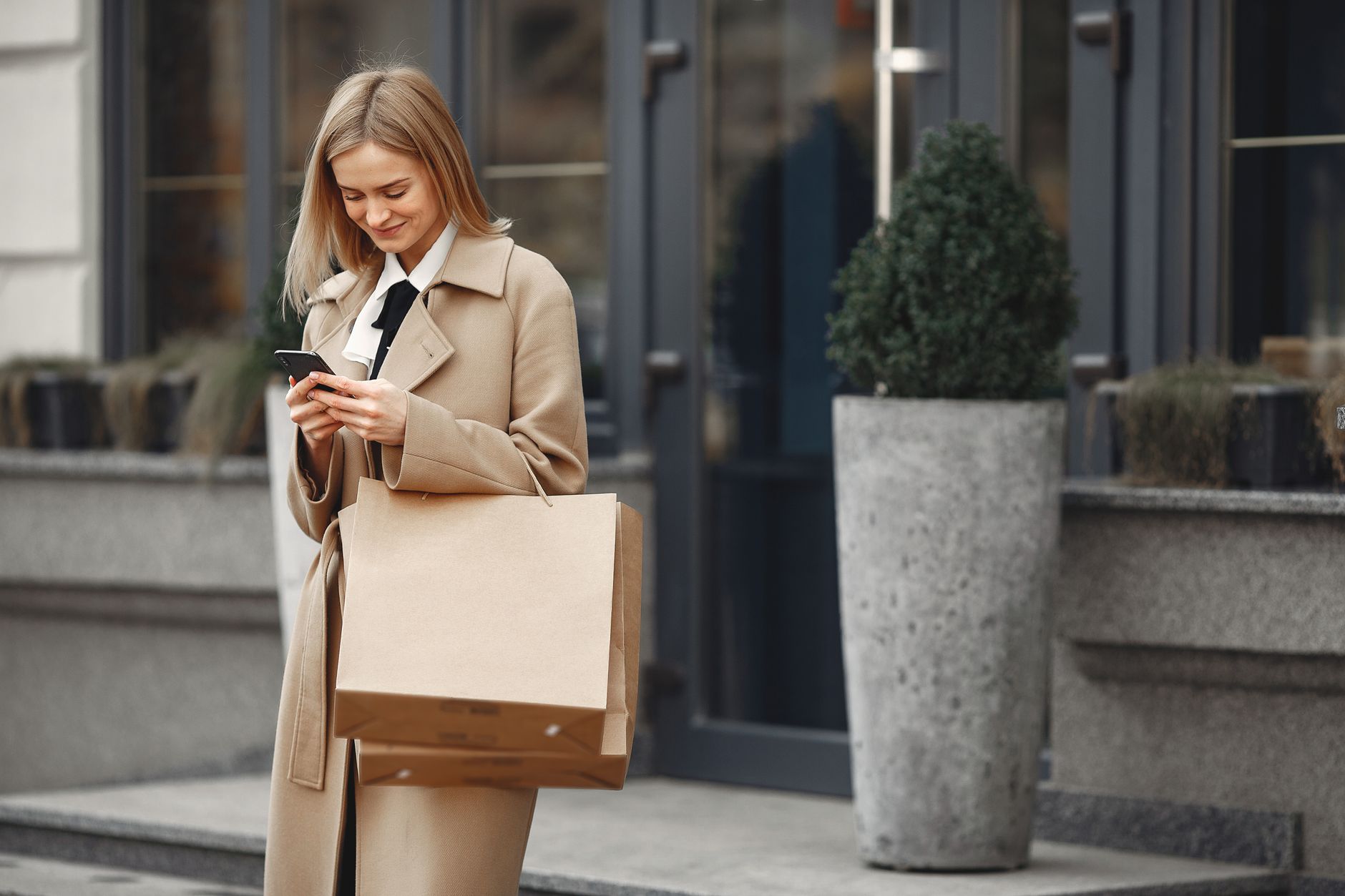 Positive elegant female in trendy coat carrying paper bags while using smartphone on street near restaurant entrance