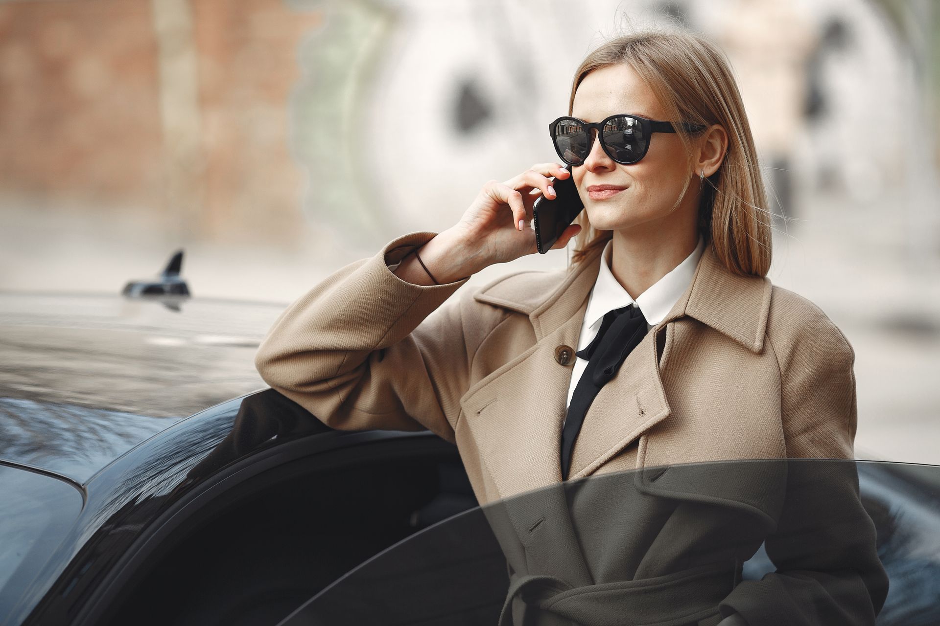 Adult female in trendy coat and sunglasses talking on smartphone leaning on modern car while standing on city street