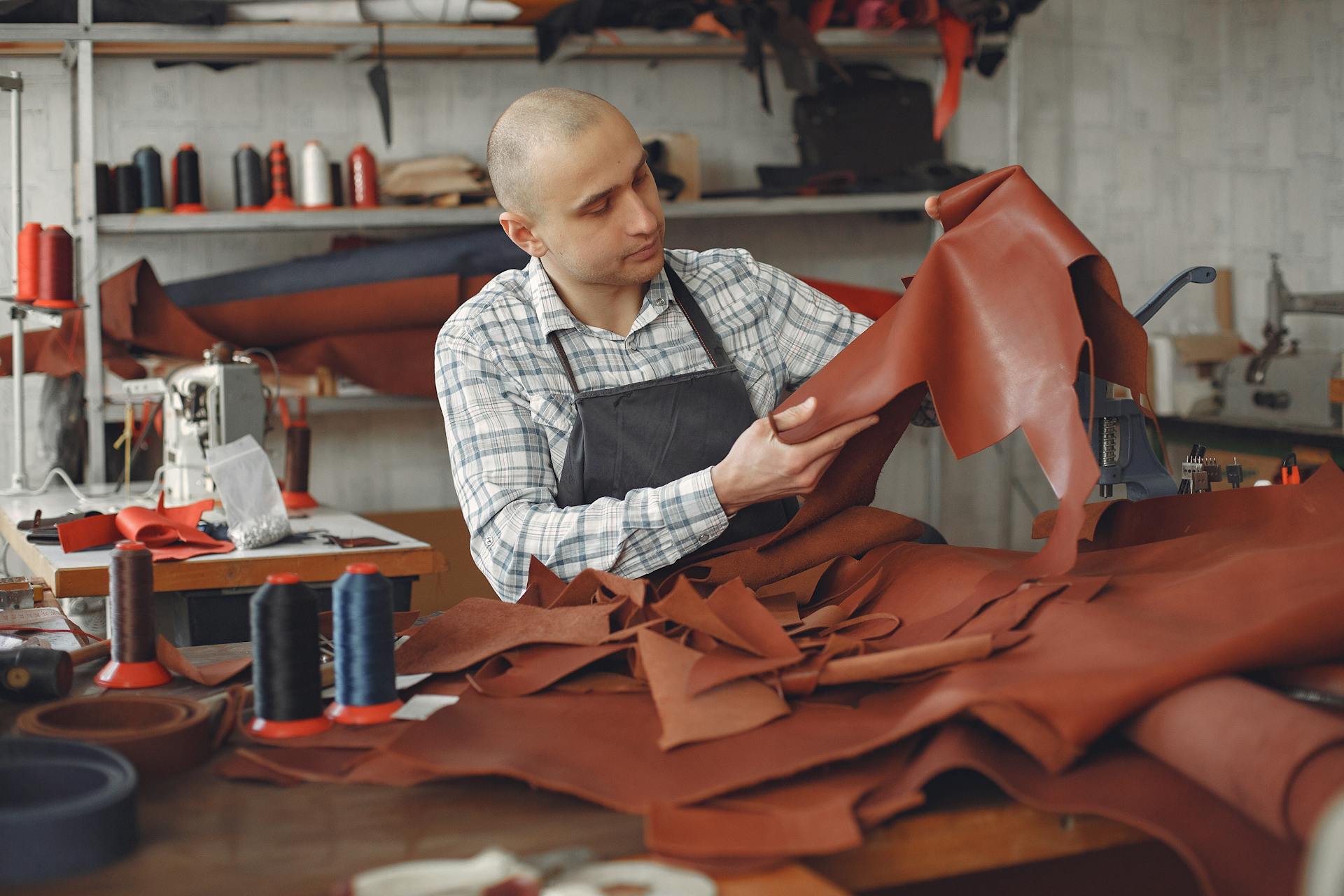 Focused adult male artisan in workwear and apron sitting in workshop among threads and cloths and sewing machines while checking leather material for quality