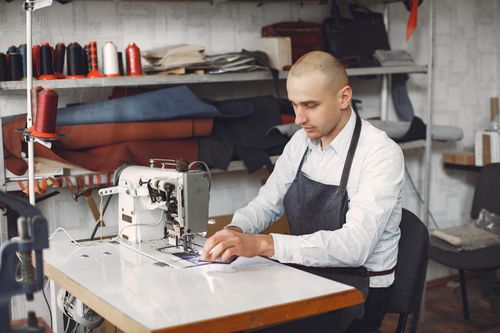 Free Side view of concentrated adult male artisan in formal clothes and apron using sewing machine while working in studio Stock Photo