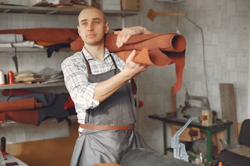 Serious focused craftsman in apron and gray check shirt standing with brown leather roll on shoulder while working in professional workshop and looking away pensively