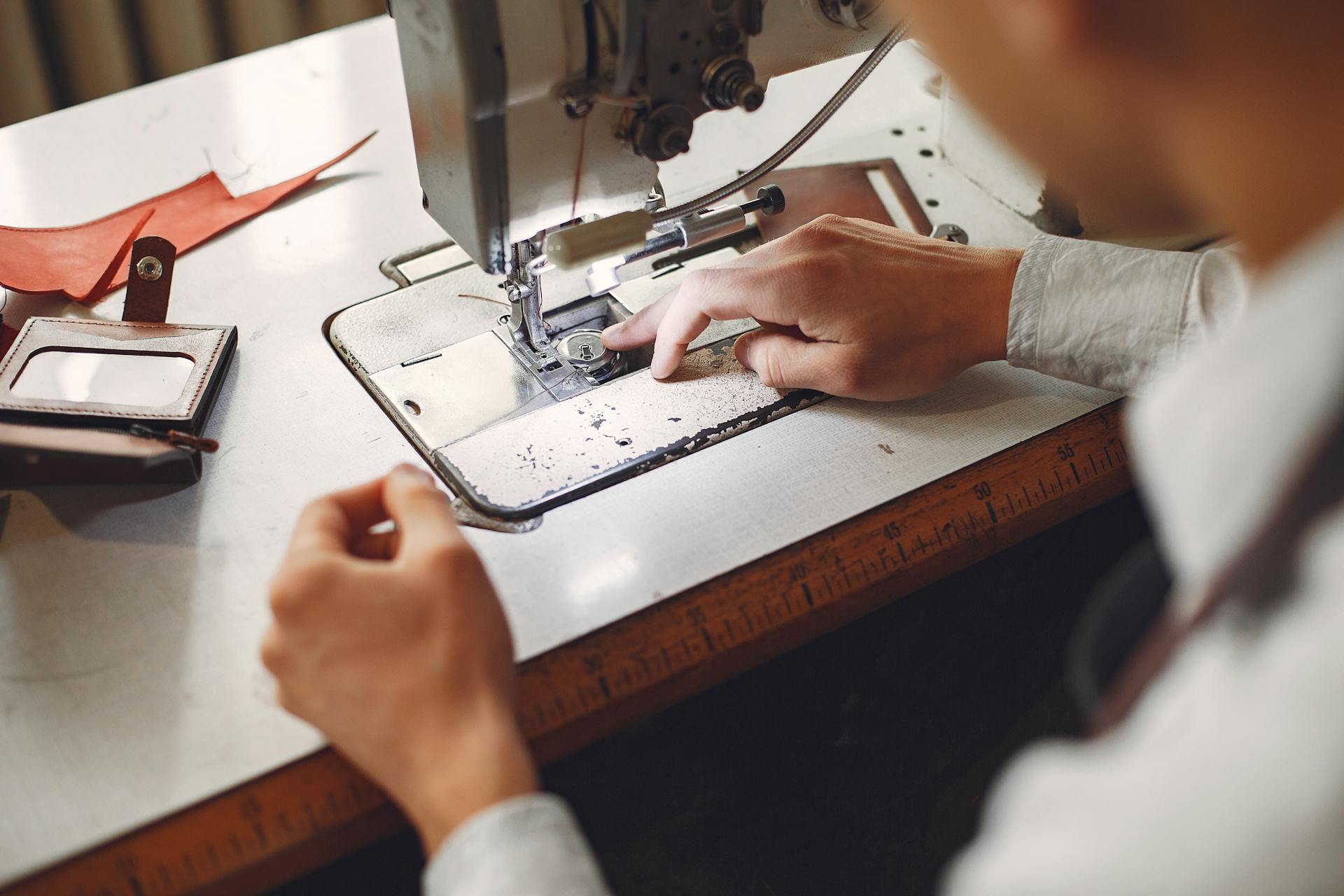 From behind crop unrecognizable artisan in white shirt sewing handmade leather details while sitting at table with sewing machine and leather wallet in daytime