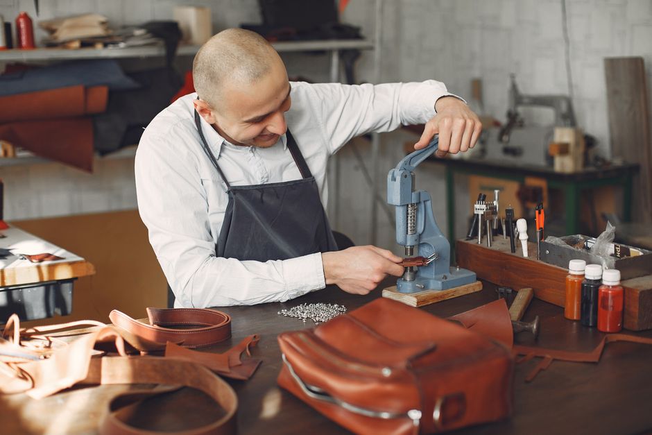 Happy young talented artisan in white shirt and apron attaching metal snaps on brown leather strap using pressing machine while working in workshop