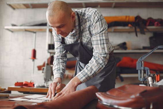 Concentrated artisan in plaid shirt and apron lining piece of leather with ruler and marker while working in professional workshop