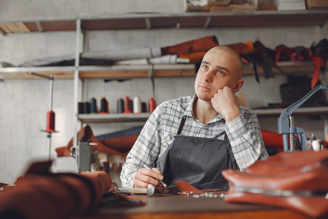 Free Positive young craftsman in check shirt and apron sitting at table in professional workshop and thinking about creating new design while leaning on hand Stock Photo