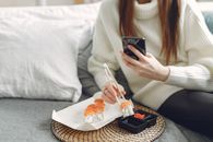 From above of unrecognizable woman in knitted sweater eating sushi with chopsticks and taking photo for social media while sitting on comfortable sofa in modern apartment