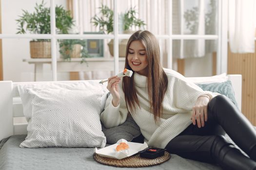 一位穿着温暖针织衫的年轻女士面带微笑，在现代客厅里，她正坐在带柔软垫子的舒适沙发上，于柳条托盘上享用新鲜的寿司