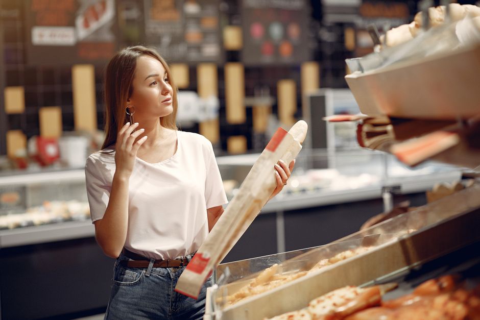 Happy young woman with long hair in casual outfit taking baguette and choosing yummy bun on showcase while visiting supermarket