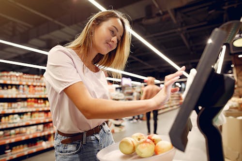 Side view of young woman in trendy clothes weighing peaches on scales while  shopping in supermarket during purchase food