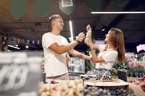 Side view of young man and woman in casual wear standing near counter and playing with each other while choosing grape in supermarket