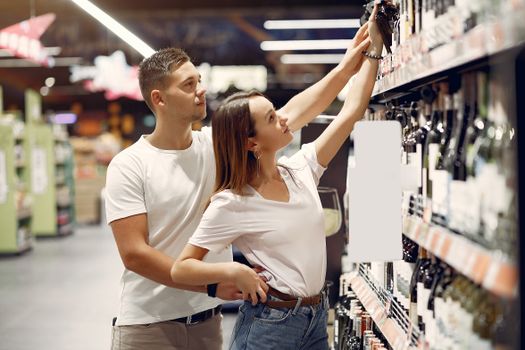 Side view of young man and woman in casual clothes standing near shelves with bottles of wine and getting bottle from top shelf together during shopping in supermarket