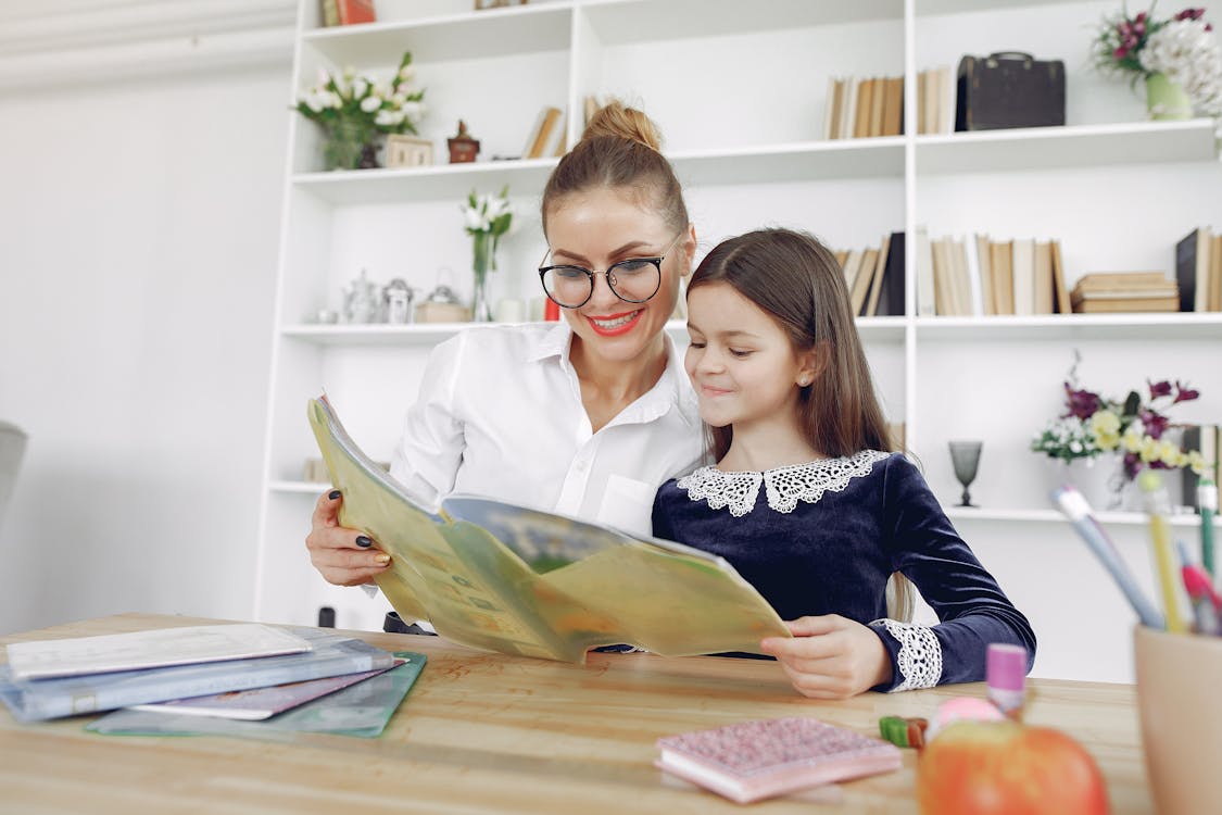 Cheerful schoolgirl and teacher reading book in library