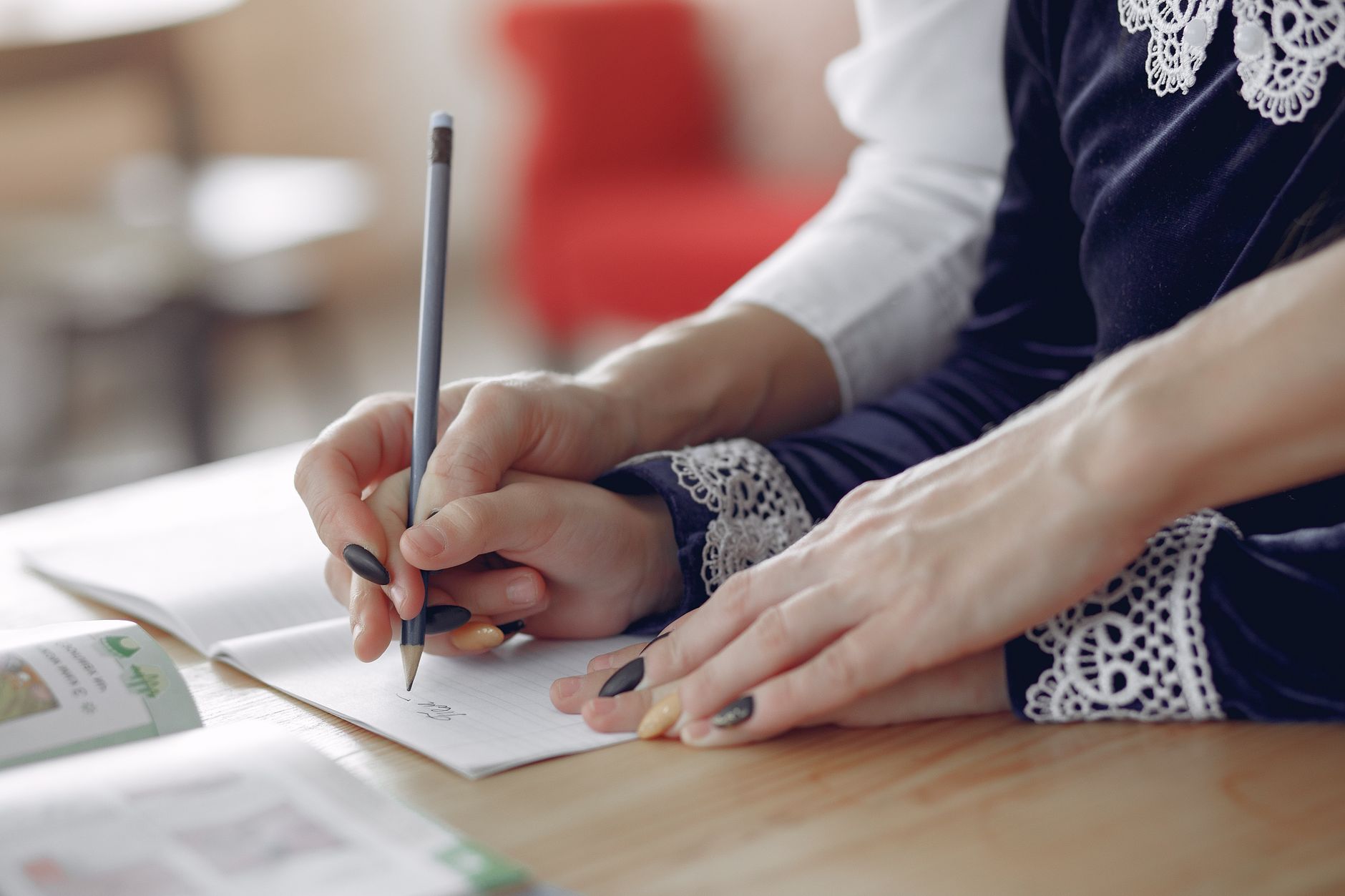 From above of unrecognizable woman holding hands girl and writing in copybook while sitting at table and working on homework in modern classroom