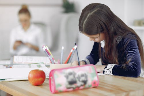 Free Focused young girl in school uniform sitting at table with stationery and apple while writing in copybook during studies against mother in modern apartment Stock Photo