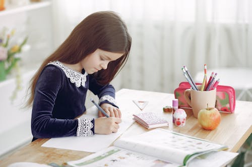 Side view of concentrated little girl sitting at wooden table at home and doing homework assignment while writing in notebook