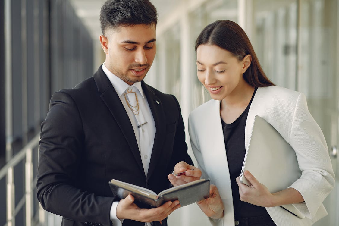 Free Focused diverse coworkers in formal clothes with notebook and laptop in hands standing in hall and discussing business project together Stock Photo