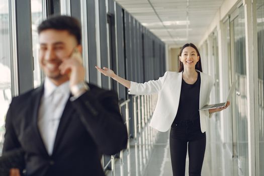 Positive female in formal outfit standing with laptop in modern office and looking at male employee talking on mobile phone