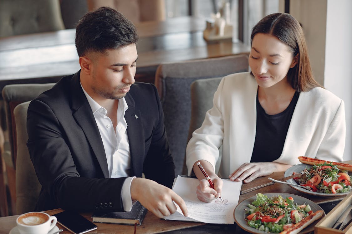Free Concentrated multiethnic colleagues in formal clothes sitting at table in cafe with cup of espresso and plates of salad while signing documents Stock Photo