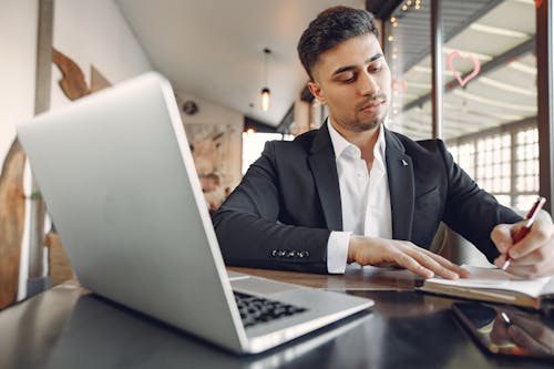 Free From below of concentrated ethnic male in formal wear sitting in creative workplace and taking notes while using laptop and working on business project Stock Photo