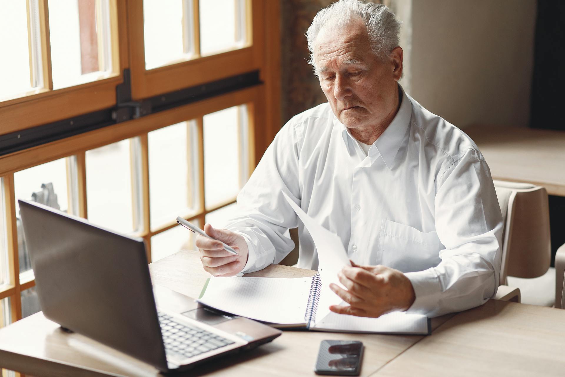 Serious gray haired aged man in white shirt sitting at table and browsing laptop while checking notes in modern office