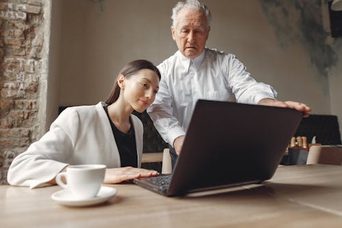 Free Low angle of positive man and woman of different ages in elegant outfit working remotely together with laptop and cup of coffee in modern workspace in loft style Stock Photo