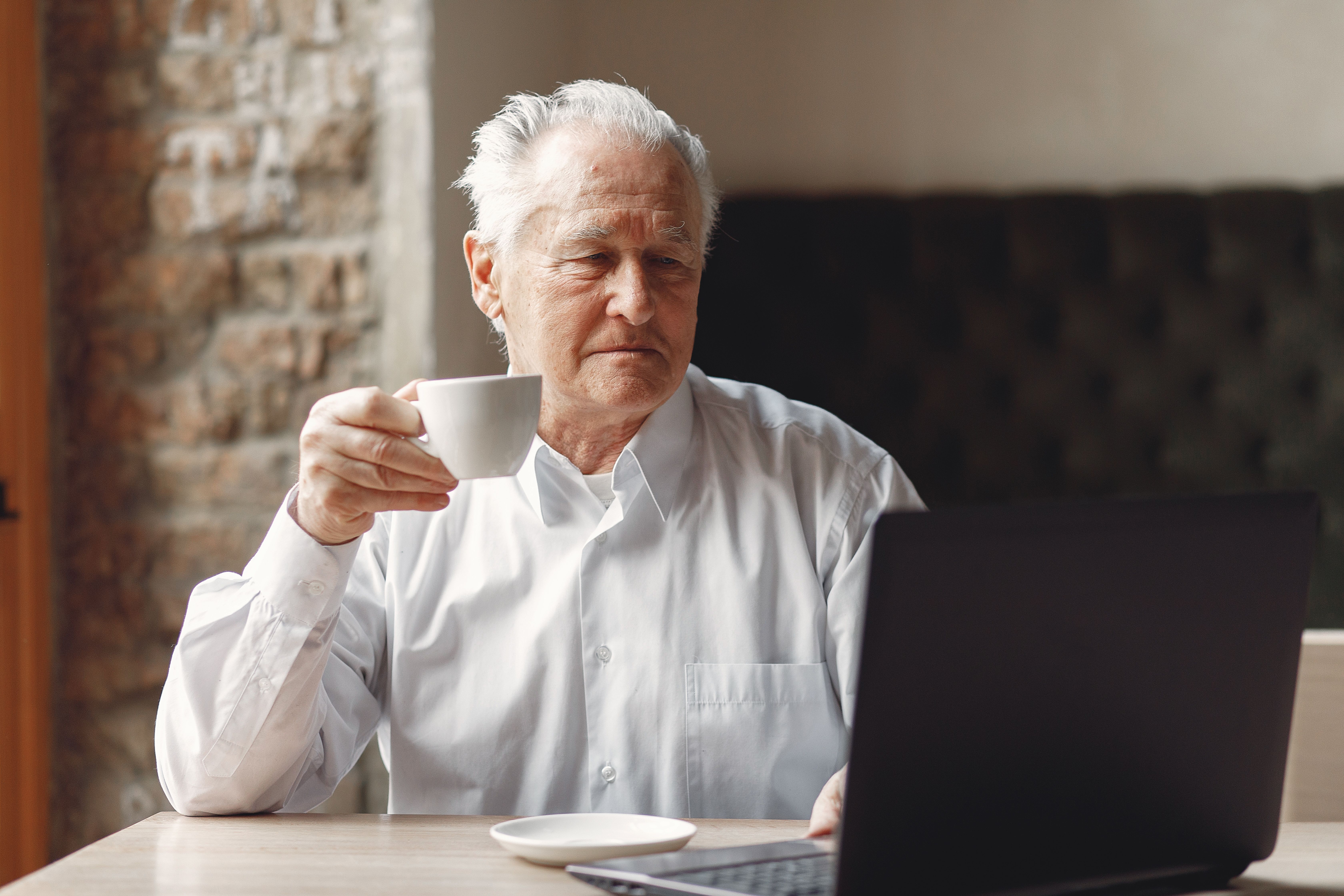 Focused elderly male enjoying cup of hot drink while surfing internet on laptop in creative modern office at daytime