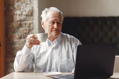 Free Focused elderly male enjoying cup of hot drink while surfing internet on laptop in creative modern office at daytime Stock Photo