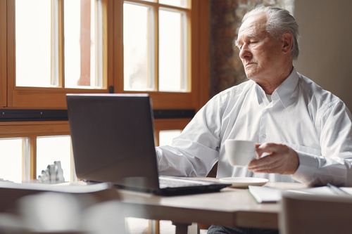 Free Relaxed aged man in smart casual outfit drinking hot drink and browsing netbook while working remotely in cafe at daytime Stock Photo