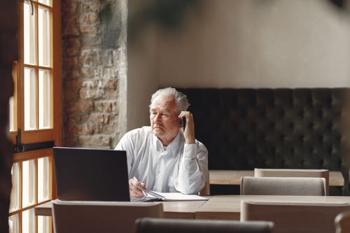 Free Thoughtfull senior male calling on smartphone and taking notes while working remotely in creative modern workspace at daytime and looking at window Stock Photo