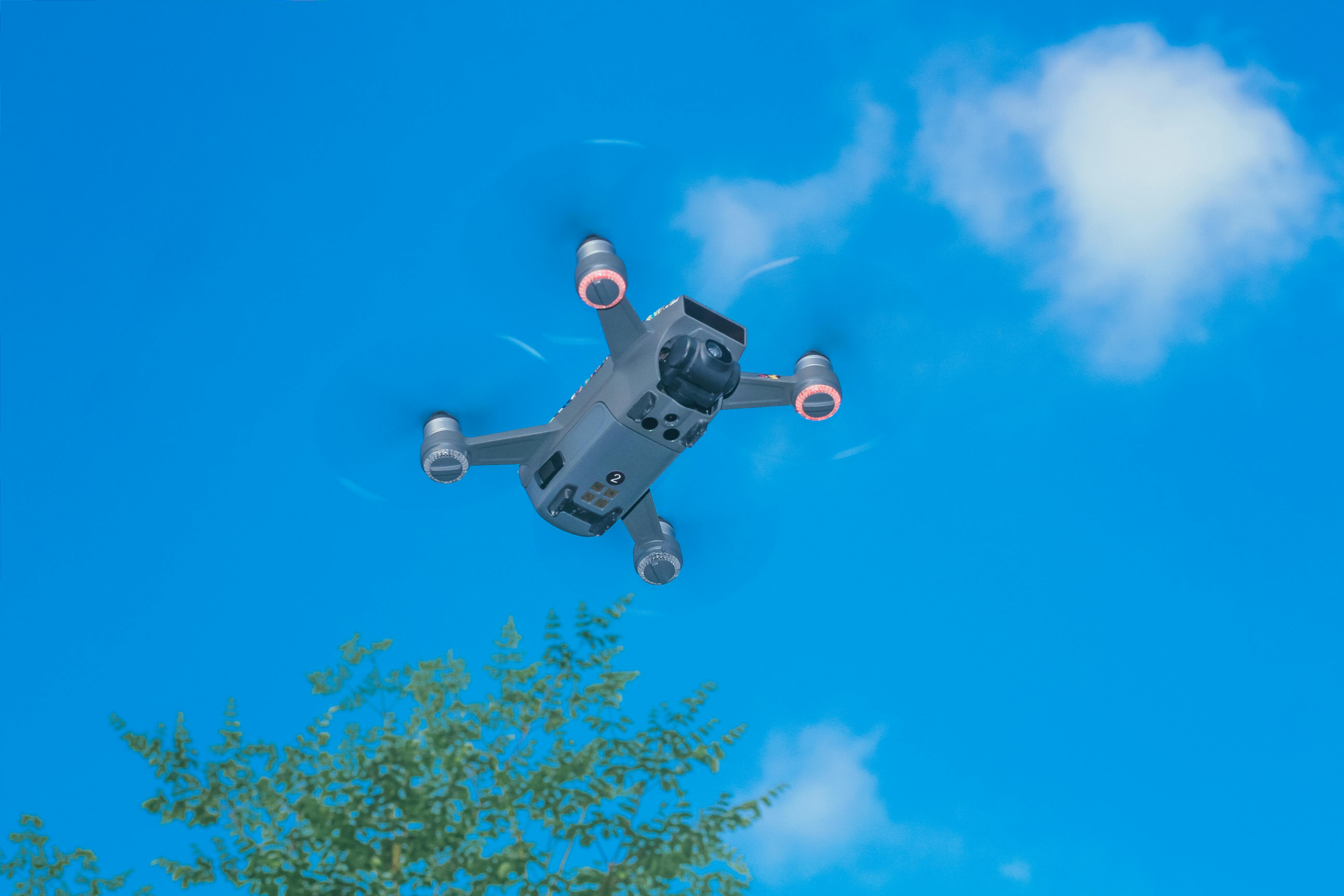 From below of modern UAV with photo camera and big propellers flying in sky above forest