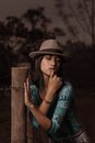Serious young woman in stylish shirt and hat touching lips and looking down while leaning hand to fence at dusk