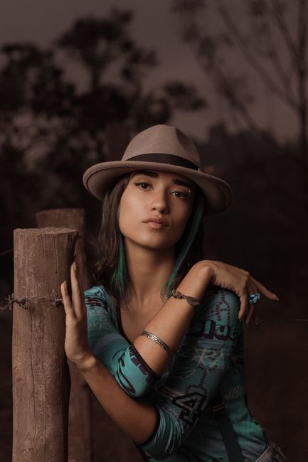Pensive young slender female in turquoise shirt and hat with makeup and accessories on wrist and arm looking at camera while leaning hand to fence at dusk