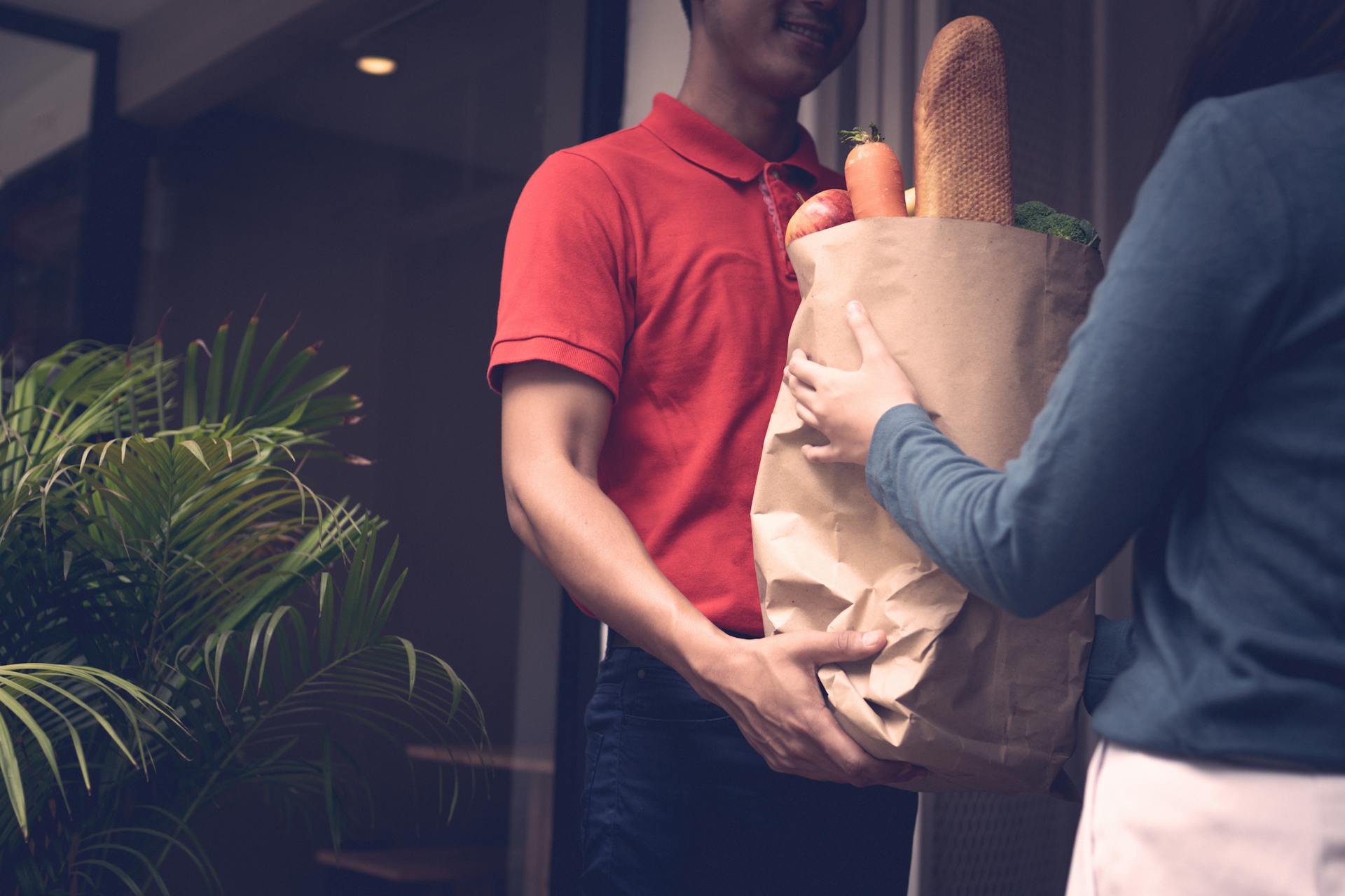 A delivery person in red shirt delivers a grocery bag filled with food items to a customer at the door.