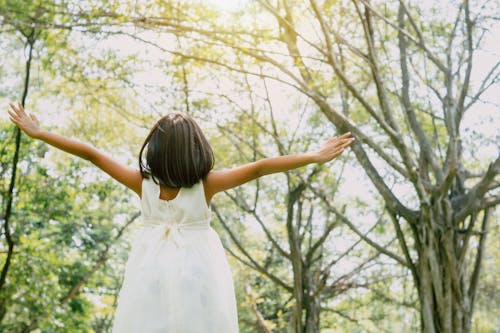 Girl in White Dress Standing Under Green Trees With Arms Outstretched