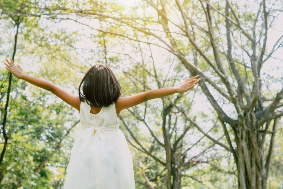 Woman in white dress with arms outstretched under sunlit forest canopy
