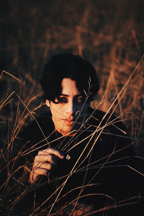 High angle of young thoughtful man wearing black clothes sitting in meadow grass