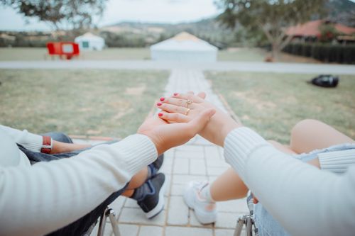 Free From above back view of crop anonymous husband holding hand of wife with manicure and golden rings while sitting on stools near narrow tiled pathway between grass lawn in countryside Stock Photo