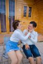 From above of young cheerful Asian lady stretching cheeks of boyfriend with grimace while looking at each other and sitting on chairs near rural wooden building and having fun together