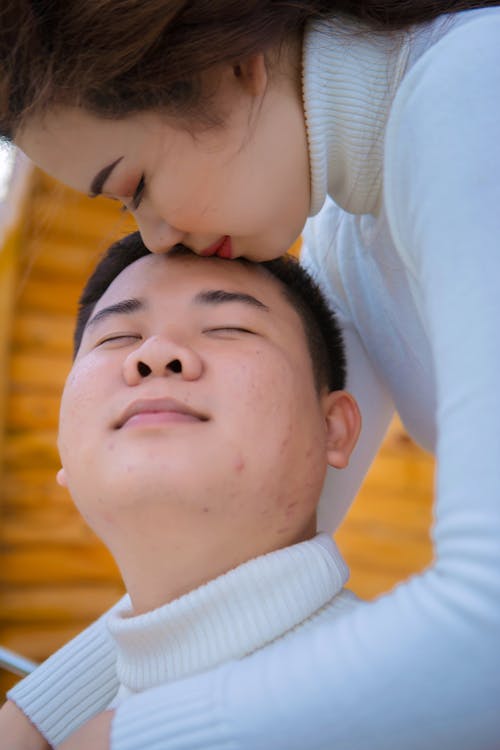 Side view of crop young ethnic female kissing gently and embracing dreamy male beloved in white knitted sweater with closed eyes while spending time together