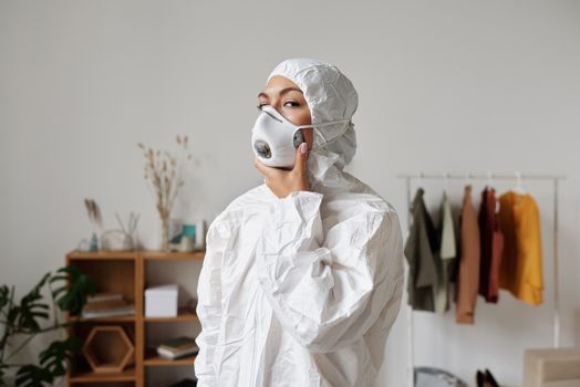 一名妇女身穿个人防护装备、戴着口罩，站在室内，周围有衣服和植物