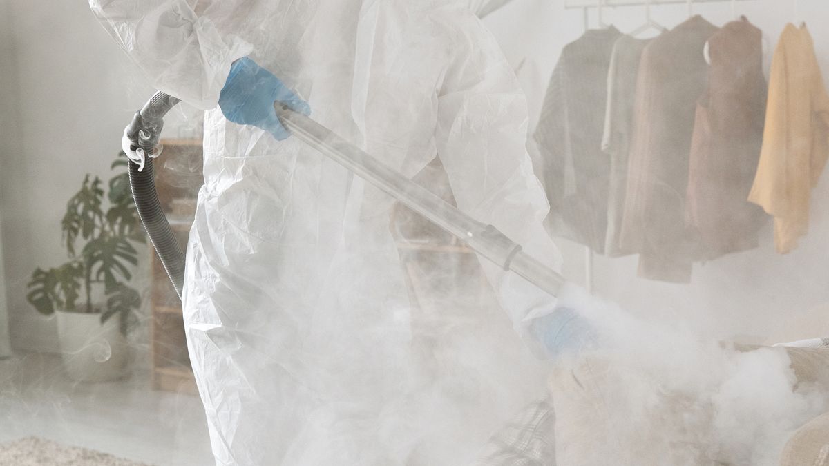 Profissional com roupa de proteção branca aplicando produto em névoa dentro de um cômodo