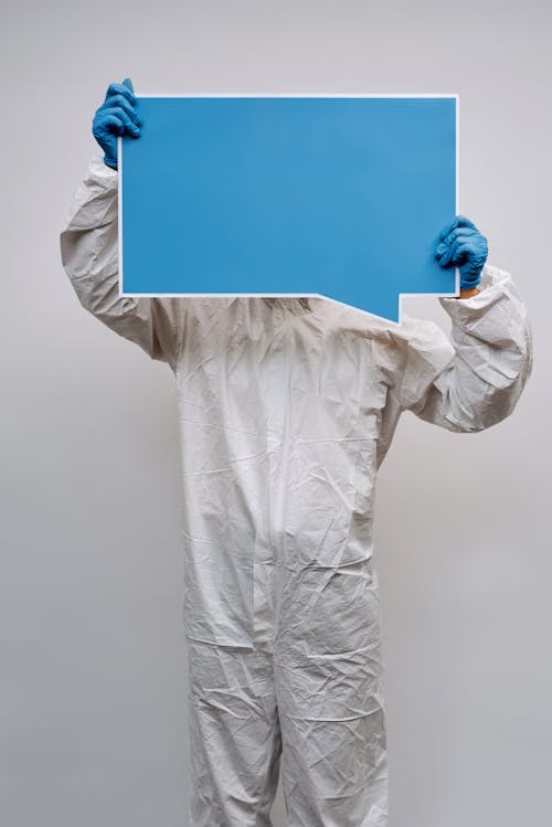 Person in White Protective Clothing and Gloves Holding a Signage Covering Face