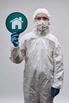Healthcare professional wearing protective suit and mask, holding a home icon sign, emphasizing safety.
