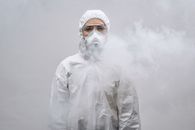 白色背景的烟雾中，站着一位穿着防护服、戴着口罩的人
