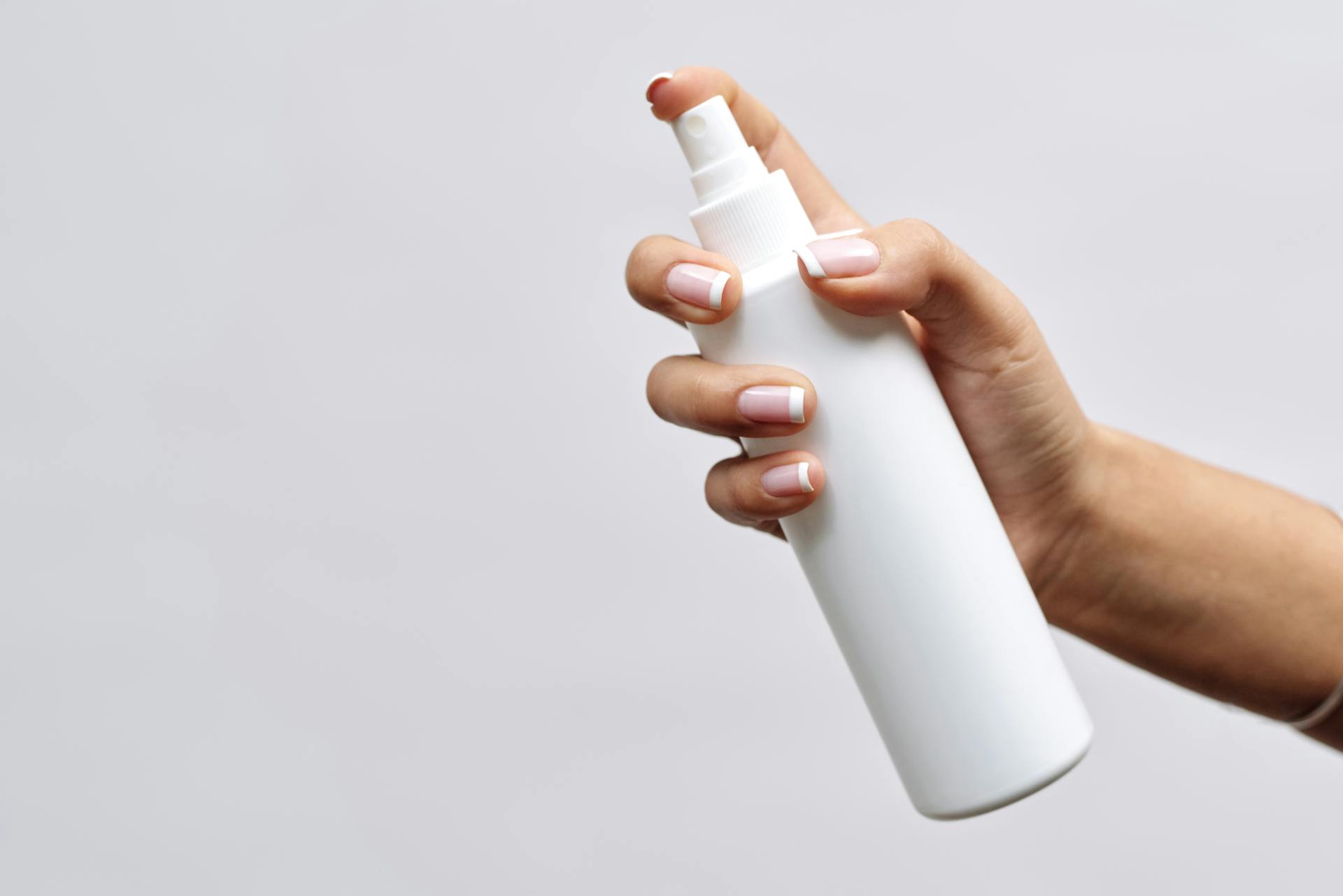 A hand with manicured nails holds a minimal white spray bottle on a white background, perfect for branding mockups.