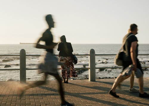 People in casual clothes walking and running on embankment while woman resting on waterfront and looking on waves during evening  on resort